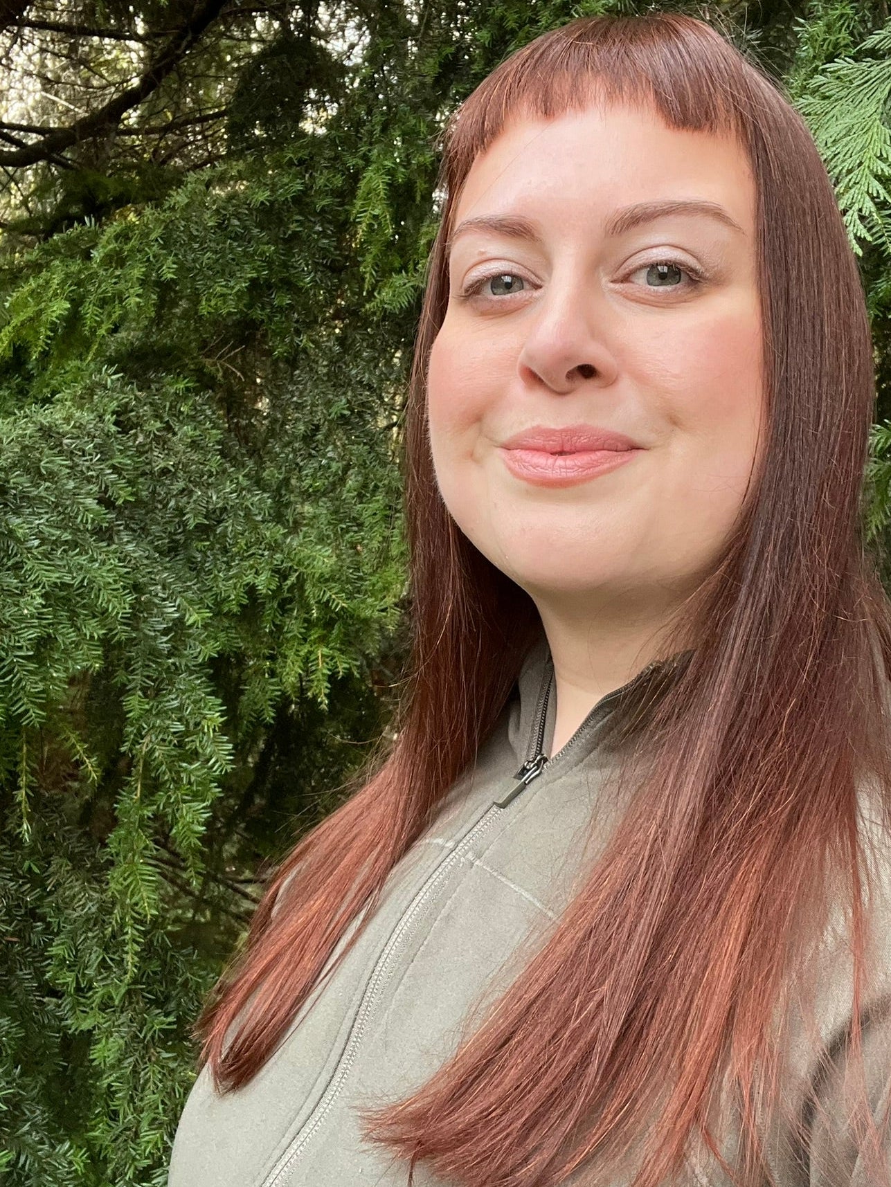 Sid Kemsley, the artist, stands in front of the forest near her Vancouver Island home. She appears with long brown hair and a sage green jacket in front of western hemlock and western red cedar foliage. 